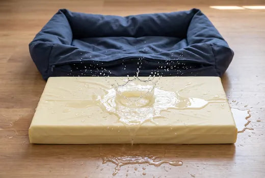 The Everyday Bed memory-foam cushion sealed in its pale-yellow waterproof liner shedding a water splash, beside the empty navy bed cover